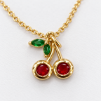 Collar dorado de cerezas rojas y hojas verdes, vista en ángulo sobre fondo blanco.